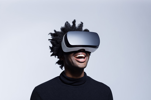 Portrait of excited afro american man wearing virtual reality glasses, standing against grey background.