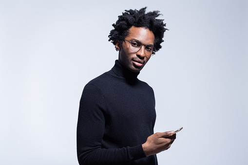 Afro american young man wearing black turtleneck and glasses holding smart phone in hand, looking at camera. Studio shot on grey background.