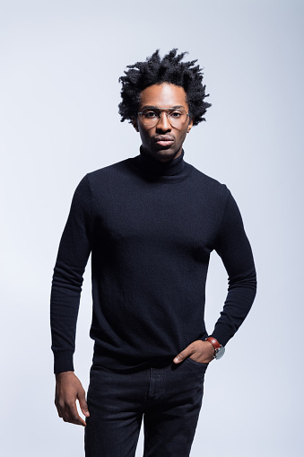 Confident afro american young man wearing black turtleneck and glasses standing with hand in pocket, looking at camera. Studio shot on grey background.