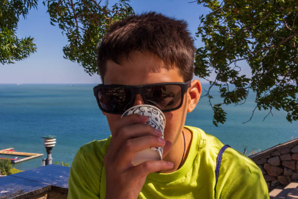 un adolescent avec des glaces foncées boit le café d’une tasse jetable sur le fond de la mer - beach on child the photos et images de collection