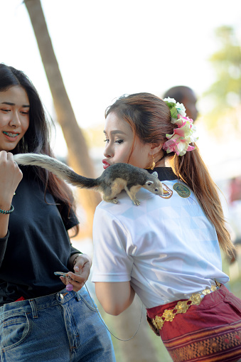 Young thai women are playing with a squirrel in Phitsanulok at daytime. One woman is traditionally dressed and is making kiss lips while squirrel is climbing on her shoulders. Other woman is watching