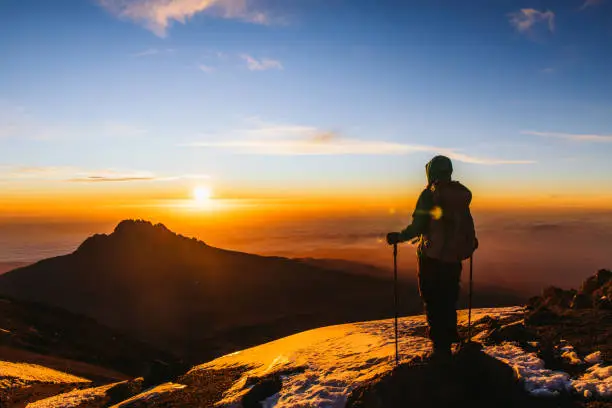 Woman with backpack and hiking poles got to the top of Africa - Mount Kilimanjaro and looking at the beautiful bright sunny sunrise above the Meru mountain