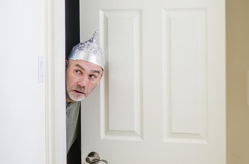 Man with aluminum foil hat hiding behind door and looking sideways with fear of electromagnetic or 5G waves