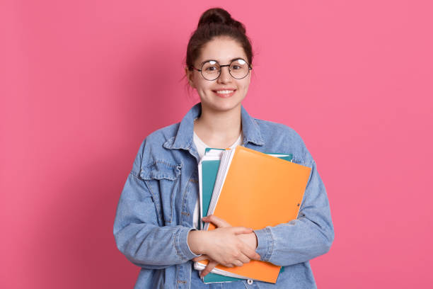 istock Friendly pleased young hipster carries colorful folder with papers, has tender smile, posing isolated over pink background, happy schoolgirl looks at camera with happy expression. 1306320937