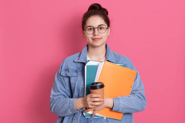 istock Young attractive student girl looking directly at camera with smile, posing isolated over rose background, wearing denim jacket and spectacles, holding paper folder and take away coffee. 1306320942