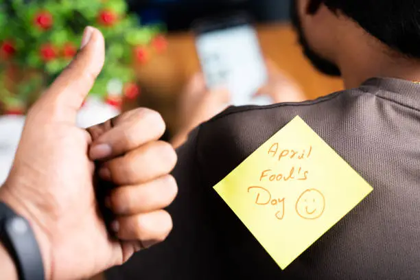 Photo of Selective focus on t-shirt, Pasting aprils fools day sticker on young mans shoulder while busy on mobile phone - concept of April fool day celebration and making fool.