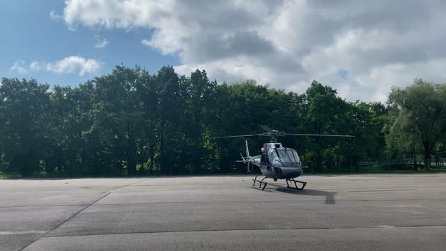 a small private helicopter on a large empty concrete take-off pad with spinning blades preparing for takeoff. trees grow in the background. large clouds are floating in the sky