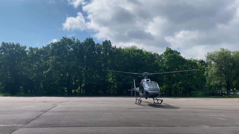 a small helicopter on a large empty concrete take-off pad with spinning blades, lifts off the ground and takes off. trees grow in the background. large clouds are floating in the sky