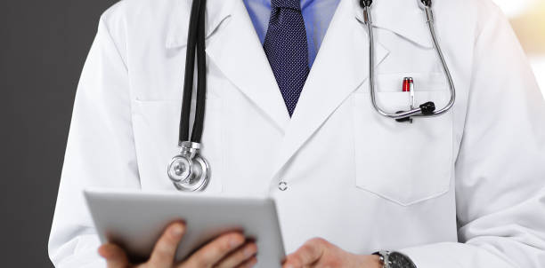 istock Unknown male doctor iusing tablet computer in his hands, while standing in sunny clinic. Medicine concept during Coronavirus pandemic. Covid-2019 1306329350