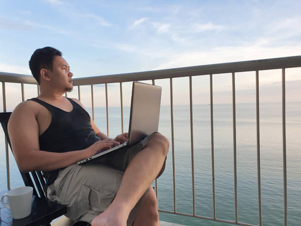 istock Happy successful Asian man using laptop while sitting on his balcony with calm blue ocean behind, remote working digital nomad 1306330350