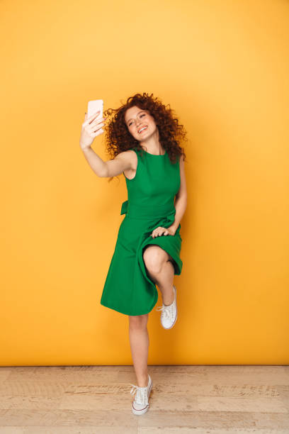 Full length portrait of a pretty redhead woman in dress Full length portrait of a pretty redhead woman in dress taking a selfie isolated over yellow background isolated color stock pictures, royalty-free photos & images