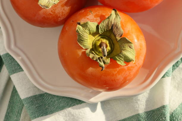 istock Fresh ripe persimmons on a white plate with a classic design on a green-white striped tablecloth, Paradise Palms 1306344764