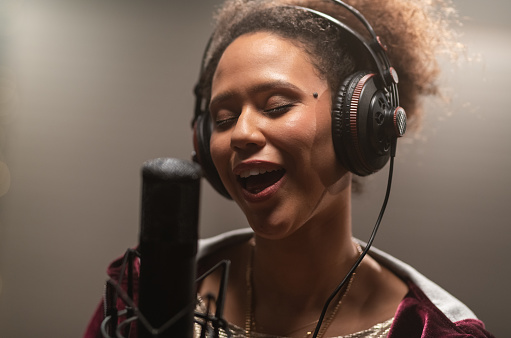 One woman, beautiful black female vocalist wearing headphones and singing into microphone in recording studio.