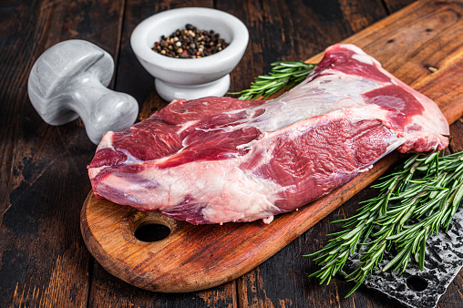 Raw lamb mutton shoulder meat on the bone on a wooden butcher cutting board with cleaver. Dark wooden background. Top view.