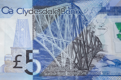 A Scottish polymer Clydesdale Bank five pound note, with an image of The Forth Rail Bridge, a cantilever bridge crossing the Firth of Forth, used for rail transportation. The blue, white and black bank note shows the numerical value of the UK currency in both numerical and written form. There is intricate detail on the note, which includes a map of Scotland, locating the site of the bridge.