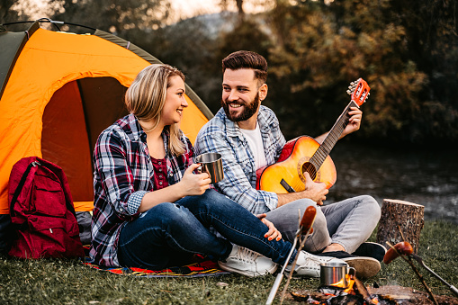 Cute heterosexual couple playing guitar and singing on camping by the river while grilling sausages on campfire.