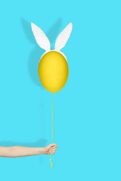 Creative easter minimal concept. Hand holding a yellow egg in bunny ears like balloon on a string on light blue background
