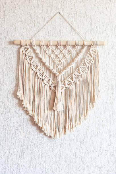 istock Handmade macrame. 100% cotton wall decoration with wooden stick hanging on a white wall.   Macrame braiding and cotton threads.  Female hobby.  ECO friendly modern knitting DIY natural decoration concept in the interior 1306383976