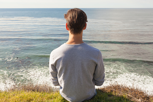 Stress-free wellness. Relaxed young man sitting alone on top a cliff facing the ocean view. Back view.