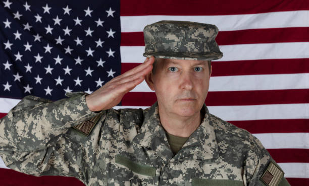 istock Man saluting while wearing military outer shirt plus cap with US flag in background 1306394747