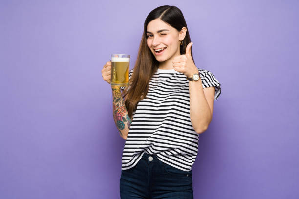Latin woman with thumbs up and drinking a beer Attractive young woman making a thumbs up and feeling happy while drinking a cold beer. Hispanic woman winking and holding a mug of beer woman drinking beer stock pictures, royalty-free photos & images