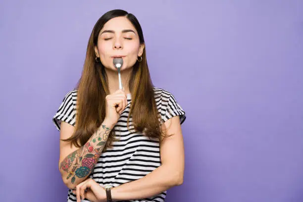 Hungry young woman with her eyes closed enjoying a spoonful of food. Pretty woman in her 20s eating and holding a spoon in her mouth