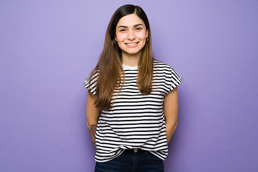 Hispanic young woman standing against a purple background with a beaming smile. Attractive woman in her 20s wearing casual clothes and looking happy