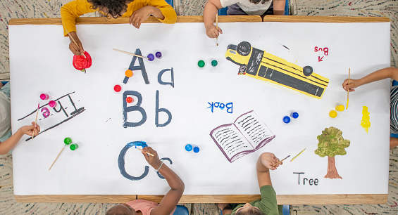 Aerial overhead view of multi-ethnic group of elementary age children coloring. The kids are seated around a table. They have coloured a school bus, book and some alphabets on the paper.