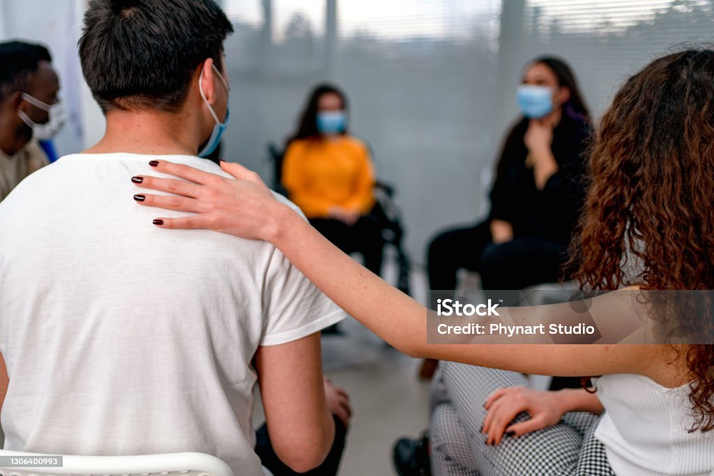 Support group patients comforting  at therapy session Support group gathering for a meeting. Young woman discussing with people at entrance hall. Males and females are sharing ideas during group therapy. Group Therapy Stock Photo