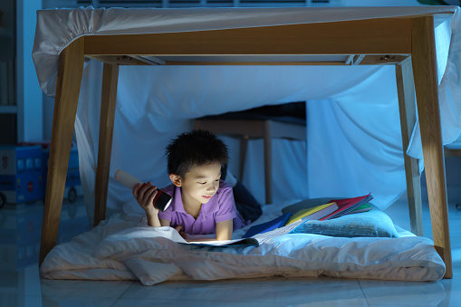 Asian preschool boy child to make a camp to play imaginatively and reading book by flashlight  in living room at home.