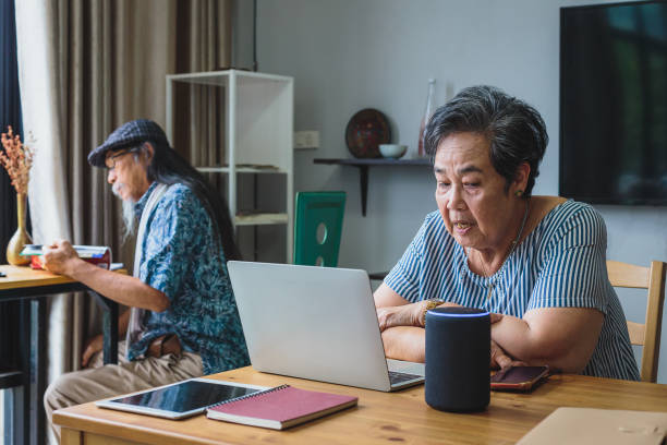 mujeres asiáticas mayores trabajando con computadora portátil y usando altavoces inteligentes mientras se instalan en la sala de estar en casa - senior couple audio fotografías e imágenes de stock
