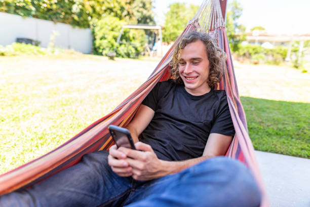 jeune homme détendant avec son téléphone dans un hamac d’arrière-cour - hamac telephone homme photos et images de collection