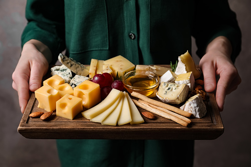 Woman holding cheese plate with honey, grissini and grapes on grey background, closeup