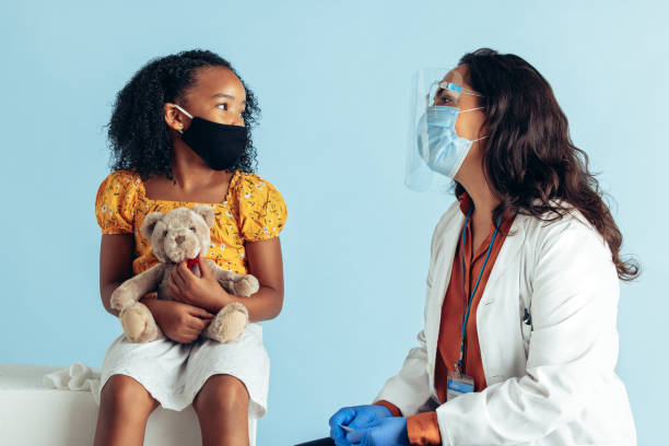 Doctor examining girl during pandemic Girl visiting doctor for check during pandemic. Female doctor wearing protective face mask and shield while examining a small girl with face mask holding her teddy bear in clinic. pediatrician stock pictures, royalty-free photos & images