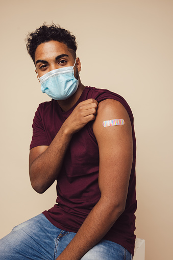 Man receiving a covid vaccine. Man wearing protective face mask showing his arm after getting vaccine shot on brown background.