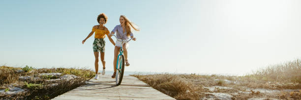 Excited woman riding bike down the boardwalk with her friends running along. Two female friends having a great time on their vacation. Panoramic shot with lots of copy space on background.