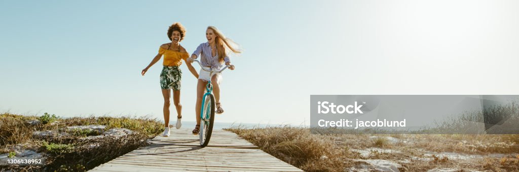 Best friends having great time on their holidays Excited woman riding bike down the boardwalk with her friends running along. Two female friends having a great time on their vacation. Panoramic shot with lots of copy space on background. Summer Stock Photo