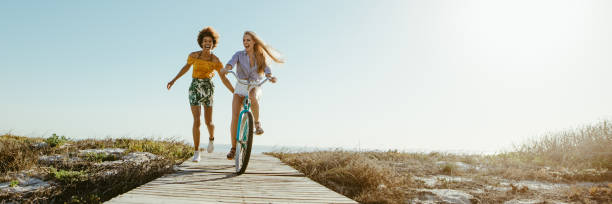 Best friends having great time on their holidays Excited woman riding bike down the boardwalk with her friends running along. Two female friends having a great time on their vacation. Panoramic shot with lots of copy space on background. summer stock pictures, royalty-free photos & images