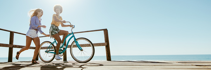 Woman riding a bicycle with her friend running behind by on boardwalk along the beach. Panorama of two female friends having fun with a bike at the seaside boardwalk.