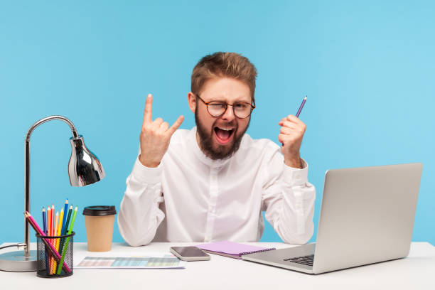 Excited positive man office worker showing rock and roll gesture sitting at workplace with laptop notepad and pencil, successfully finished annual report