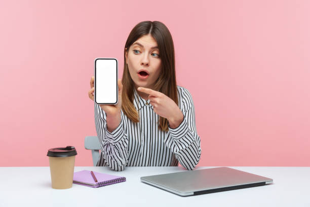 istock Shocked surprised woman office worker pointing finger at smartphone with white empty display, looking with astonishment sitting at workplace 1306450367