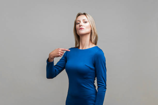 This is me! Portrait of arrogant businesswoman pointing at herself. indoor studio shot isolated on gray background This is me! Portrait of arrogant businesswoman in tight elegant blue dress pointing at herself, looking egoistic and haughty, proud of achievement. indoor studio shot isolated on gray background egocentric stock pictures, royalty-free photos & images