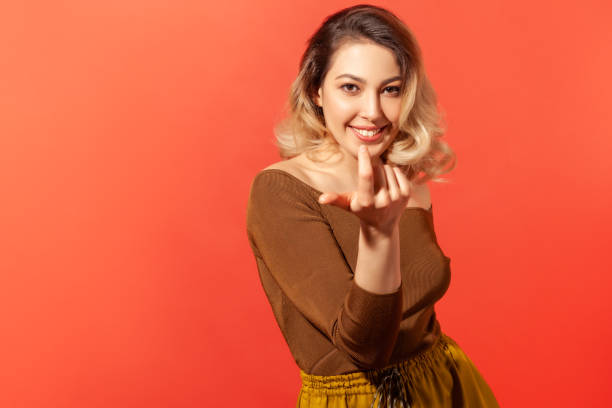 Woman calling with one finger, making beckoning gesture Come here, follow me! Portrait of pretty blonde haired woman looking playfully and calling with one finger, making beckoning gesture, inviting to come. Indoor studio shot isolated on red background beckoning stock pictures, royalty-free photos & images