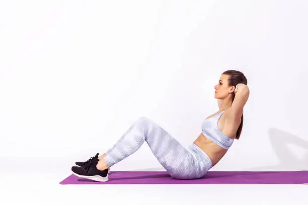 Full length side view athletic woman in white tights and top lying on mat and training doing sit ups, exercising abdominal muscles, lower-body workouts. Indoor studio shot isolated on gray background