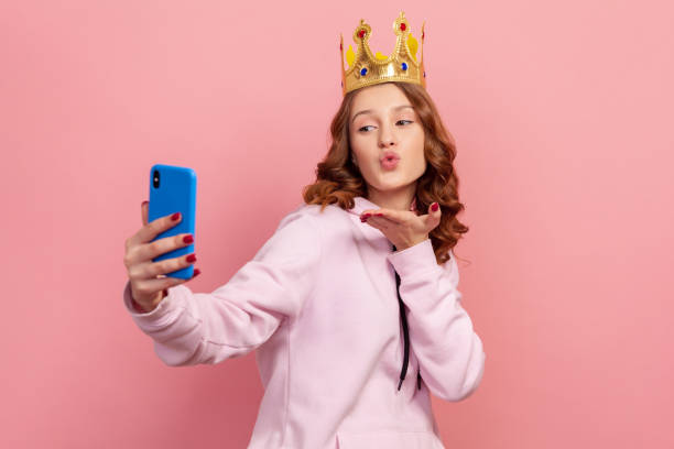 Portrait of confident curly haired teenage girl in hoodie with crown sending air kiss on cellphone camera, modern technology, selfie Portrait of confident curly haired teenage girl in hoodie with crown sending air kiss on cellphone camera, modern technology, selfie. Indoor studio shot isolated on pink background queen royal person photos stock pictures, royalty-free photos & images