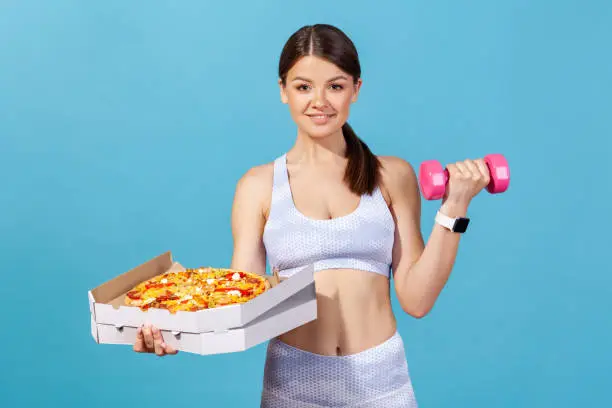 Active sportswoman in white tights and top pumping muscles with dumbbell holding pizza box, keeping diet, rejecting from junk food. Indoor studio shot isolated on blue background
