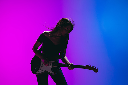 Solo performance. Silhouette of young Caucasian girl, female guitarist posing isolated over gradient blue-pink background in neon light. Concept of music, art, human emotions. Copy space for ad.