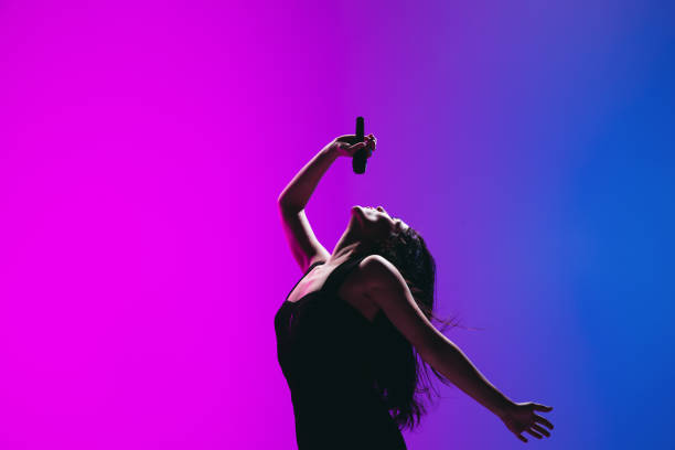 Portrait of young female singer isolated on blue-pink background. Half-length portrait of young girl, female singer, musician with microphone isolated on gradient blue-pink background in neon light. Concept of music, art, show, performance. Copy space for ad singer stock pictures, royalty-free photos & images