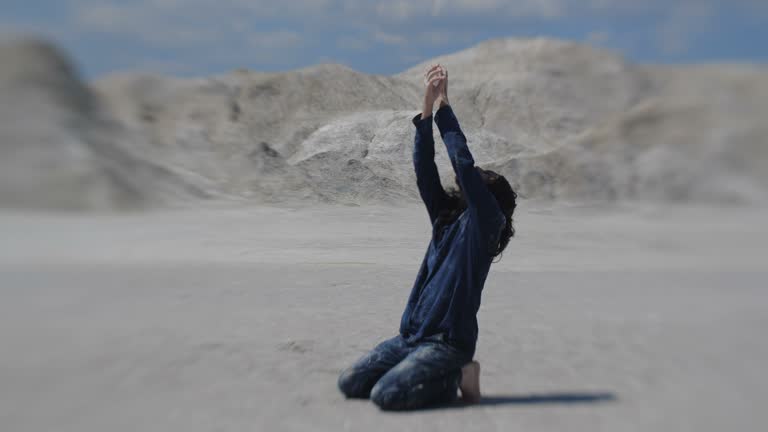 Lonely exhausted traveler goes mad in  desert. Long-haired man in dirty clothes dancing crazy dance on sand kneeling and raising hands 4k slow motion 50 fps