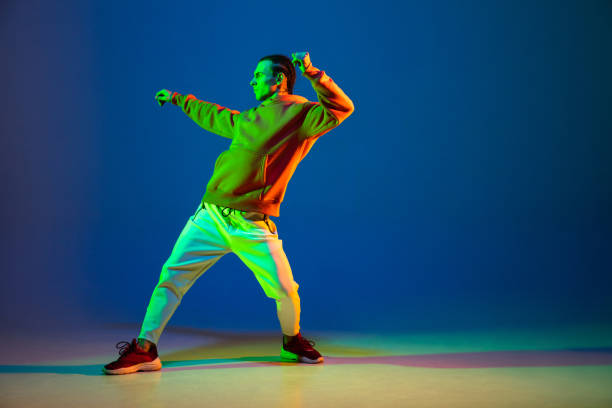 Young man dancing hip-hop in stylish attire on blue background in neon light Street style. Hip-hop male dancer athletic man dancing in stylish attire over colorful background at studio in blue neon light. Youth culture, fashion, action, healthy lifestyle. Copy space for ad. fluorescent photos stock pictures, royalty-free photos & images